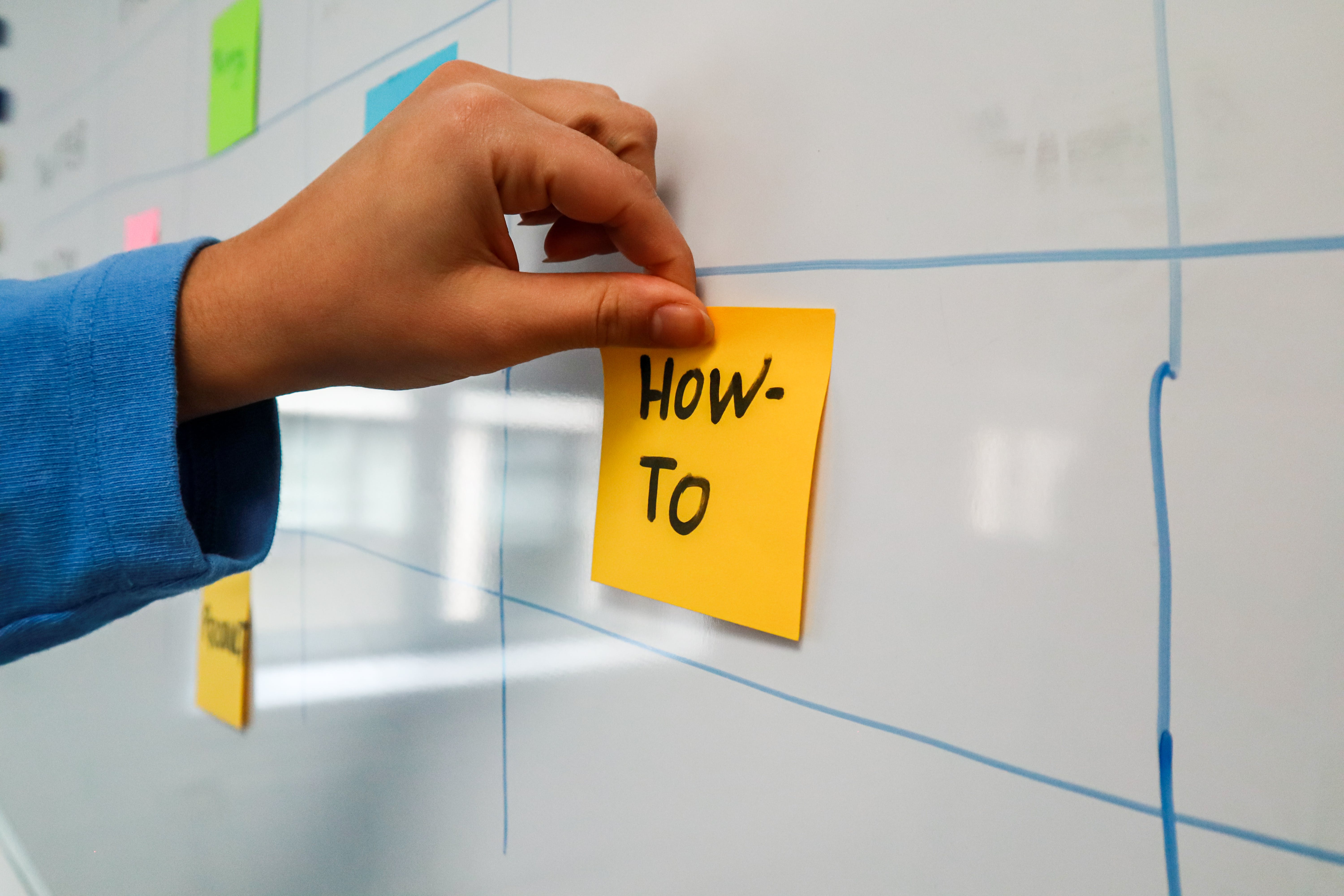 Hand posting a sticky note to a wall stating "How To"
